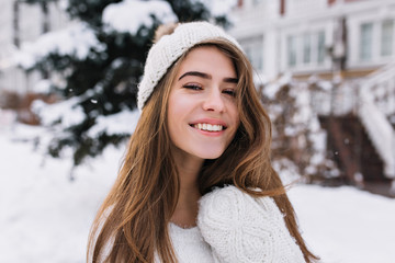 Close-up portrait of pleased blonde woman with sincere smile enjoying winter morning. Lovely european girl in white hat looking at snowy view outdoor..