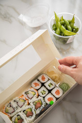 Frau isst Edamame, Maki und Inside Out California Sushi vom Lieferdienst mit Lachs, Surimi, Avocado, Gurken mit Ingwer, Wasabi und Soja Soße in Einweg Bento Box Verpackung