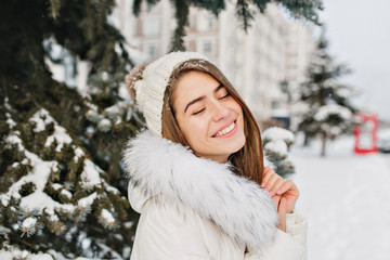 Close up portrait of amazing joyful girl in white warm warm clothes enjoying winter time in city. Young pretty woman in snow smiling with closed eyes. Happy emotions, christmas coming, new year mood