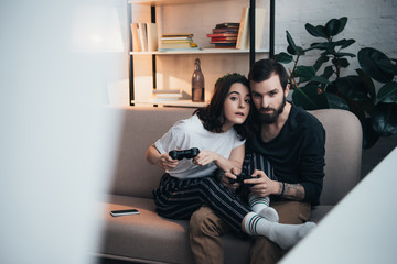 Fototapeta premium beautiful young couple sitting on couch with joysticks and playing video game in living room with copy space