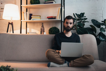 handsome bearded man sitting on couch, looking at camera and using laptop in living room