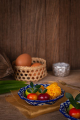 Thai desserts on a plate of white and blue stripes placed on golden cloth and wood table there are pandan, water bowl, egg in basket, Similar object, fork and coconut placed around.
