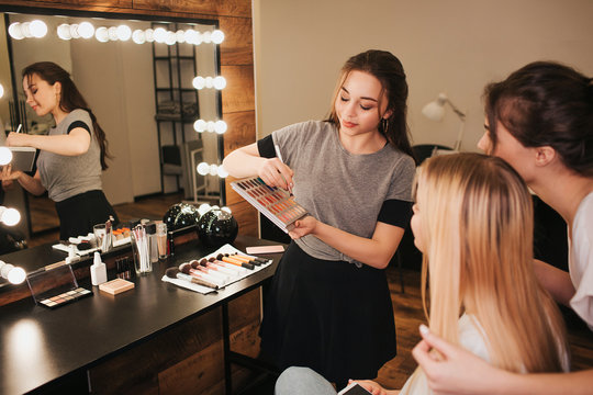 Young professional make up artists with client in room. Woman on left hold eyeshadows palette and brush. Blonde and bruette women look in mirror together.