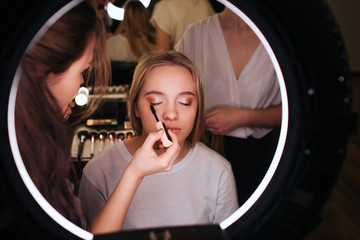 Look in mirror in beauty room. Make up artist put mascara on eyelashes. Young blonde woman sit on chair with closed eyes. Hardresser work behind client.