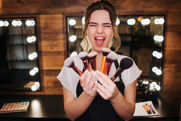 Emotional young make up artist stand in beauty room and scream. She hold set of brushes in hands. Mirror with light bulbs behind.