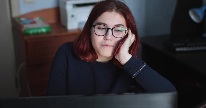 European girl in glasses speaks on the phone near the computer