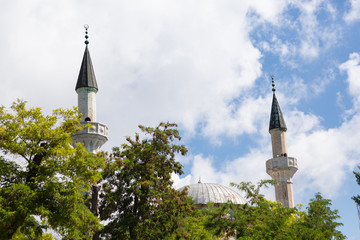 Mosque dome and two minarets protrude from the trees, against a blue sky with clouds, Muslim...