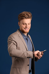 Fashionable young man with phone looking at watch on color background