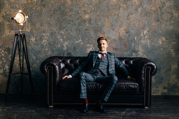 Young handsome man posing for a fashion shoot in a studio. Fashion as a lifestyle. Model sitting on a sofa in tuxedo. Successful fashionable businessman. Business look advertising