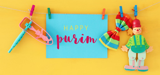 Purim celebration concept (jewish carnival holiday) with note, clown and noisemaker over wooden yellow background