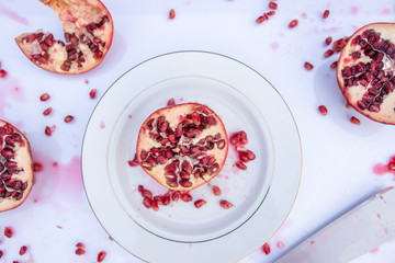 Juicy Pomegranate Fruit cut with seeds and juice on white background. Ceramic plate and knife. Top down.