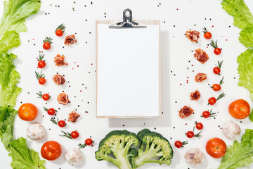 top view of empty clipboard among tomatoes, lettuce leaves, prosciutto, broccoli, spices and garlic