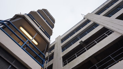 Looking up at a brutalist car park in nottingham with blue railings, halogen lights and a large exterior staircase towards the roof on an overcast day with no sunshine