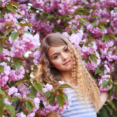 Adorable young girl with curly hair in pink dress walking in blossom cherry garden in sunny spring day