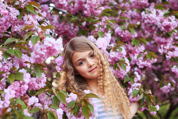 Fototapeta premium Adorable young girl with curly hair in pink dress walking in blossom cherry garden in sunny spring day