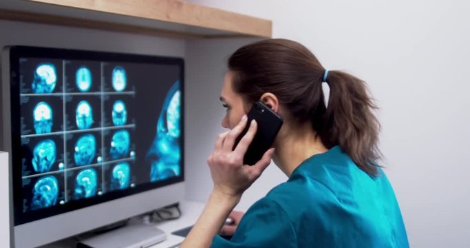 Professional female doctor neurologist talking on the phone while examining brain MRI scans on a computer screen at her personal desk at hospital. 4K UHD