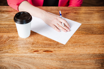 A woman's hand holds a pen over a sheet of paper and a glass of coffee on a wooden table.