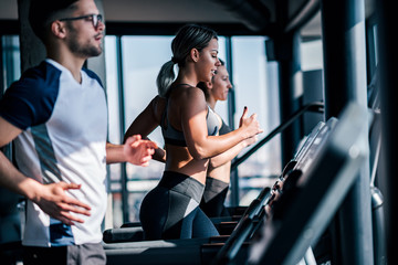 Side view of fit young man and women running side by side on treadmills at the gym.