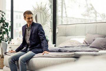 Handsome cheerful gentlemen having seat on bed in furniture showroom