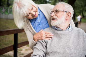 smiling senior woman looking at happy husband in wheelchair, while holding hand on his shoulder