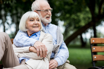 smiling senior man embracing happy wife while sitting together on bench in park
