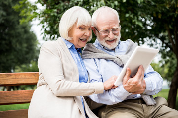 happy smiling senior couple using digital tablet while sitting on bench in park - obrazy, fototapety, plakaty