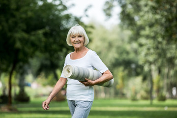 happy senior woman with fitness mat walking in park