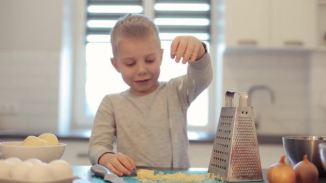 Little beautiful caucasian boy with big blue eyes cooking in the bright kitchen. He is pours cheese. Cheese grater. Happy cheerful kid make pizza.