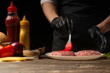 The cook rubs beef burgers with butter, against the background with ingredients, a restaurant business, fast food, tasty food, gastronomy, cooking, menu. Horizontal photo