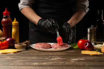 The cook rubs beef burgers with butter, against the background with ingredients, a restaurant business, fast food, tasty food, gastronomy, cooking, menu. Horizontal photo