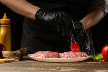 The cook rubs beef burgers with butter, against the background with ingredients, a restaurant business, fast food, tasty food, gastronomy, cooking, menu. Horizontal photo
