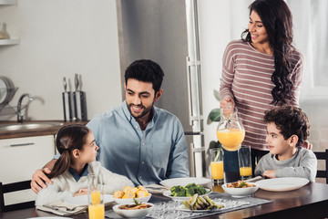 cheerful latin mother holding jar with orange juice near son while looking at husband hugging daughter during lunch - Powered by Adobe