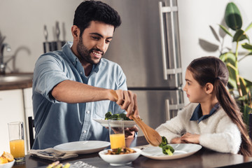Fototapeta na wymiar cheerful latin father putting broccoli in plate of cute daughter at home