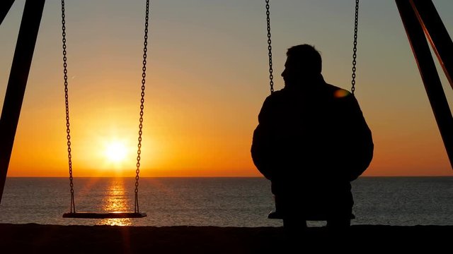 Back view silhouette of a sad man complaining alone missing his partner siting on swing at sunrise