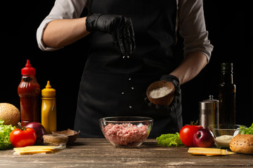 Chef salting minced meat to create a burger patty. Against the background with ingredients for a...