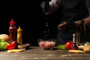 Chef salting minced meat to create a burger patty. Against the background with ingredients for a burger. Gastronomy, recipes, menus, fast food. Juicy hamburger