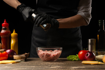 The chef peppers the fresh ground beef for the patties in the burger. On the background are the ingredients for the burger. Horizontal photo. Cooking delicious food