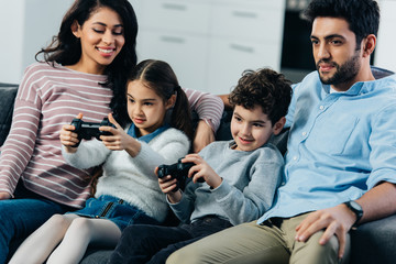Fototapeta premium cheerful parents sitting on sofa near kids playing video game at home