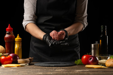 The chef prepares the cutlet with fresh ground beef, holding in the form of a heart, ingredients on the background, cooking a delicious burger. Recipe book, homemade food, menu, cooking