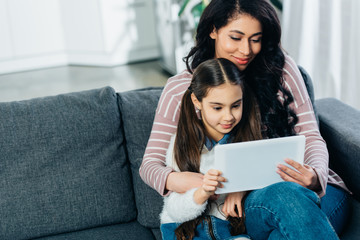 latin woman sitting on sofa with daughter and using digital tablet at home