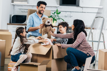 cheerful latin father holding soft toy near hispanic family while unpacking boxes in new home