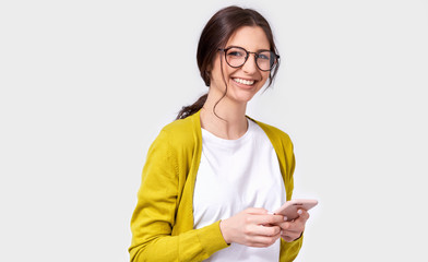 European beautiful brunette smiling young woman using cellphone, messaging to her friend. Happy female chatting with her boyfriend via smart phone, looking at the camera. People, technology, emotion