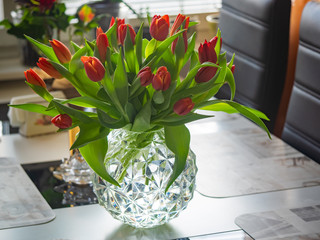 Glass vase with red tulips flowers in light cozy interior. White table and light from the open window