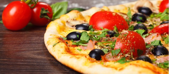 Close up Italian pizza with melted cheese, red cherry tomatoes, black olives, ham, and green leaves on a brown table decorated with mushrooms, red hot chili pepper and cherry tomatoes