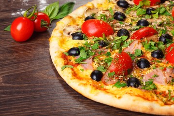 Close up Italian pizza with melted cheese, red cherry tomatoes, black olives, ham, and green leaves on a brown table decorated with mushrooms, red hot chili pepper and cherry tomatoes
