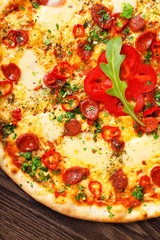 Close up Italian pizza with melted cheese, sausage, snack sticks, pepper and fresh green oregano leaves on a brown table decorated by mushrooms, red sweet pepper and cherry tomatoes