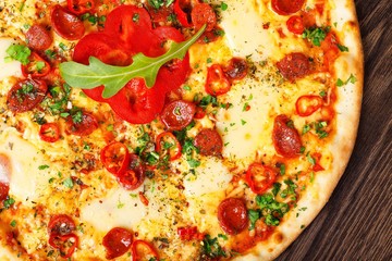 Close up Italian pizza with melted cheese, sausage, snack sticks, pepper and fresh green oregano leaves on a brown table decorated by mushrooms, red sweet pepper and cherry tomatoes