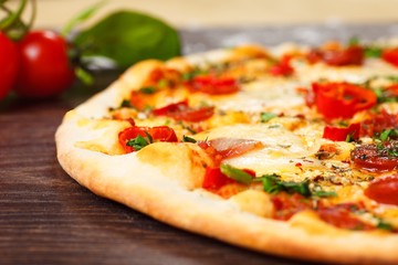 Close up Italian pizza with melted cheese, sausage, snack sticks, pepper and fresh green oregano leaves on a brown table decorated by mushrooms, red sweet pepper and cherry tomatoes