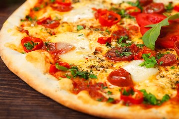 Close up Italian pizza with melted cheese, sausage, snack sticks, pepper and fresh green oregano leaves on a brown table decorated by mushrooms, red sweet pepper and cherry tomatoes
