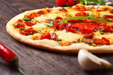 Close up Italian pizza with melted cheese, sausage, snack sticks, pepper and fresh green oregano leaves on a brown table decorated by mushrooms, red sweet pepper and cherry tomatoes